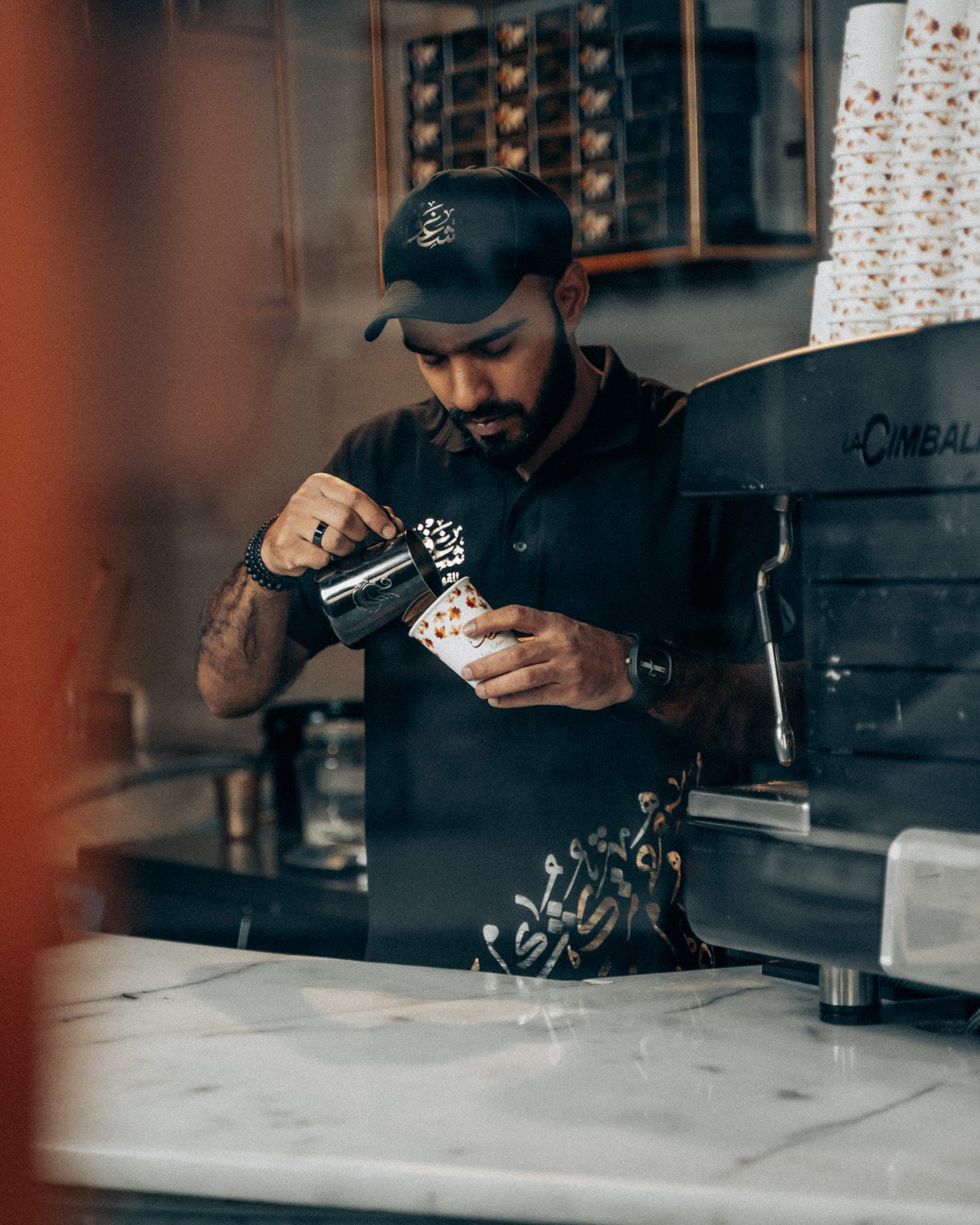 A Shaghf barista pouring latte art into a paper cup at the espresso machine